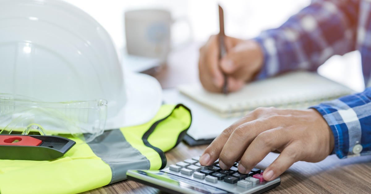 A person using a calculator and writing in a notebook beside a white hard hat, safety glasses, and a high-visibility vest.
